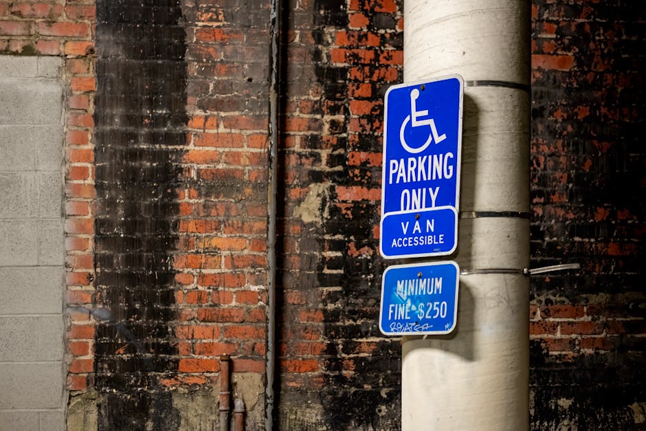 A tall, light grey concrete utility pole standing against a weathered red and black brick wall with visible mortar and signs of aging. Two blue and white parking restriction signs are attached to the pole: the upper sign indicates parking is reserved for individuals with disabilities, with a wheelchair symbol and text reading 'Parking Only,' and an additional smaller sign below states 'Van Accessible.' The lower sign specifies a minimum fine of $250 for violations. The scene is set outdoors, with shadows indicating daylight, and the pole and signs are positioned near a narrow urban street or alleyway, which may be relevant for planning house removals and furniture transport in Harringay. The image does not show any vehicles or moving activities but provides context for parking regulations relevant to home relocation or moving services, as exemplified by Movers Harringay.