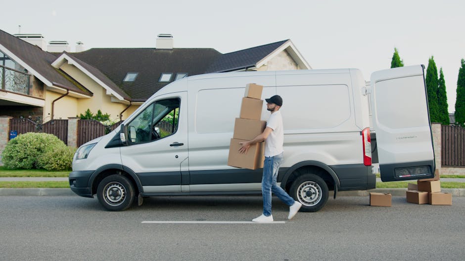 A man wearing a white t-shirt, jeans, white sneakers, and a black cap is loading cardboard boxes into the side door of a white panel van parked on a residential street. The attic ladder of the van is open, and additional cardboard boxes are placed on the pavement nearby. In the background, there are modern detached houses with pitched roofs, dormer windows, a wooden fence, and well-maintained greenery including bushes and tall trees. The scene appears to be part of a home relocation or furniture transport process, with the man actively engaged in packing and moving materials as part of a professional removals service. The environment is well-lit with natural daylight, emphasizing the organized loading process typical of house removals managed by Movers Harringay.