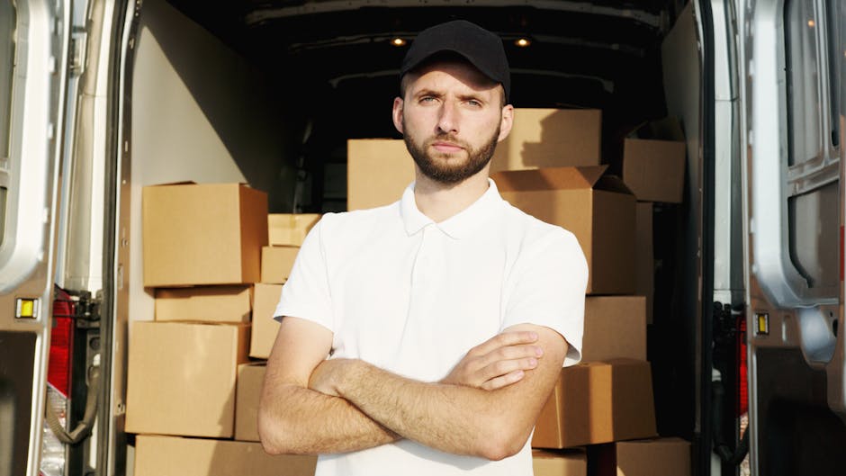 A young man with a short beard and dark hair, wearing a white polo shirt and a black cap, stands inside an open moving van filled with stacked brown cardboard boxes and packing materials. The boxes are arranged neatly, some wrapped in plastic or protective padding, indicating a home relocation process. The interior of the van is dark with a visible floor and walls designed for transporting furniture and belongings. The man is crossing his arms and looking directly at the camera with a serious expression, suggesting concentration during the loading or unloading process. The van is parked on a paved area, and natural lighting illuminates the scene, emphasizing the organized packing and preparation for furniture transport. This image is relevant to house removals and moving services offered by Movers Harringay, highlighting the logistics involved in packing and loading for a residential move.