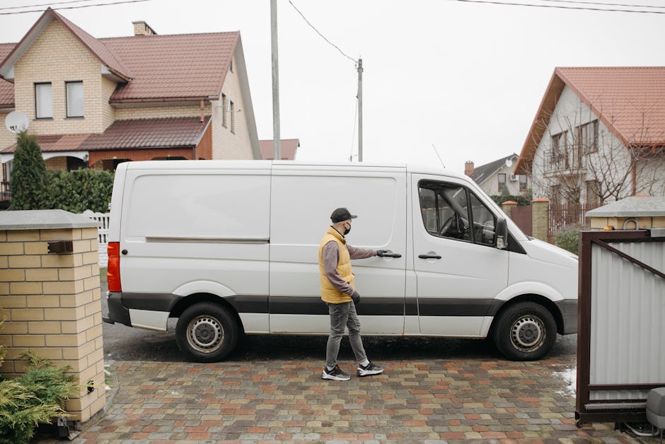 A man wearing a white t-shirt, jeans, white sneakers, and a black cap is loading cardboard boxes into the side door of a white panel van parked on a residential street. The attic ladder of the van is open, and additional cardboard boxes are placed on the pavement nearby. In the background, there are modern detached houses with pitched roofs, dormer windows, a wooden fence, and well-maintained greenery including bushes and tall trees. The scene appears to be part of a home relocation or furniture transport process, with the man actively engaged in packing and moving materials as part of a professional removals service. The environment is well-lit with natural daylight, emphasizing the organized loading process typical of house removals managed by Movers Harringay.