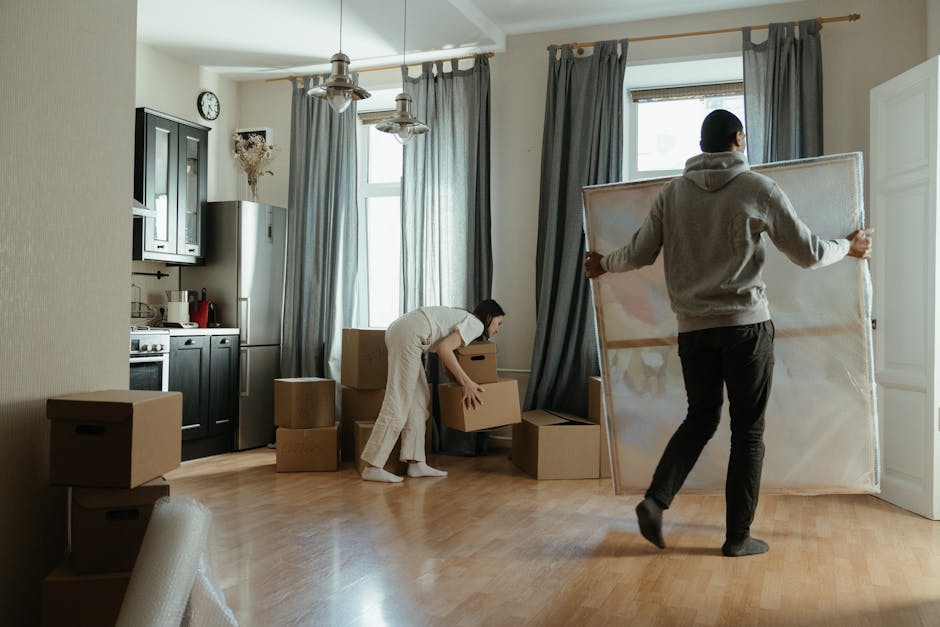 Inside a residential property, two movers are actively engaged in the home relocation process as part of a furniture transport project. The room features light wooden flooring, with natural light entering through two large windows draped with dark grey curtains. One mover, dressed in a hoodie and dark trousers, is carrying a framed mirror or painting, holding it upright by its edges. The other mover, wearing a white top and beige trousers, is bending down to pick up a cardboard box, which is part of a collection of packed boxes of various sizes distributed across the floor and nearby furniture. Several other cardboard boxes are stacked or positioned near the kitchen area, which includes a black cabinet, a silver refrigerator, and a stove with appliances on top, all situated against a beige wall. There are also hanging pendant lights, and a glimpse of a clock on the wall, indicating the ongoing packing and loading process typical of house removals, carried out by professionals such as Movers Harringay.