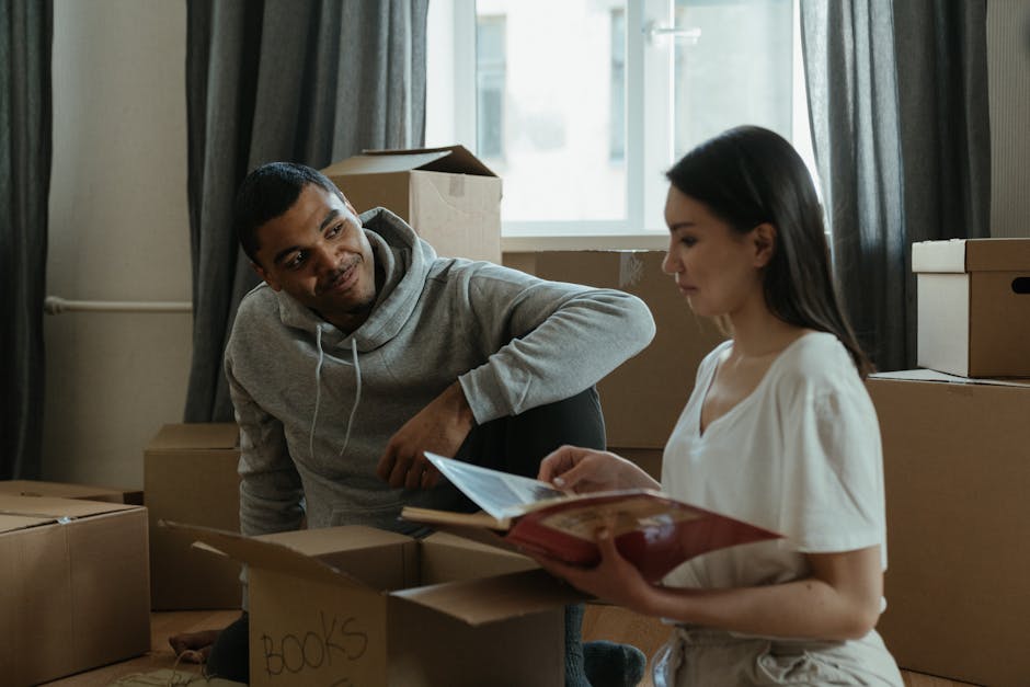 Inside a residential property, two movers are actively engaged in the home relocation process as part of a furniture transport project. The room features light wooden flooring, with natural light entering through two large windows draped with dark grey curtains. One mover, dressed in a hoodie and dark trousers, is carrying a framed mirror or painting, holding it upright by its edges. The other mover, wearing a white top and beige trousers, is bending down to pick up a cardboard box, which is part of a collection of packed boxes of various sizes distributed across the floor and nearby furniture. Several other cardboard boxes are stacked or positioned near the kitchen area, which includes a black cabinet, a silver refrigerator, and a stove with appliances on top, all situated against a beige wall. There are also hanging pendant lights, and a glimpse of a clock on the wall, indicating the ongoing packing and loading process typical of house removals, carried out by professionals such as Movers Harringay.