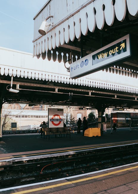 A view of Golders Green Underground Station with its elevated canopy featuring striped fabric awnings and white trim, under which several individuals are seen standing and sitting on wooden benches. Some figures are engaged in lifting or carrying cardboard boxes and duffle bags, indicating the process of home relocation or moving services. The scene captures a loading zone adjacent to the train tracks, with a yellow plastic container or trolley positioned nearby for transporting belongings. Beneath the canopy, an overhead sign directs towards the exit and indicates the location of lifts, while a CCTV camera is mounted on the station’s structure. Sunlight filters through, illuminating the area and highlighting the practical aspects of packing, loading, and transporting household or office items as part of a furniture transport or residential move, with the actions aligning with the services provided by Movers Harringay in the context of house removals.