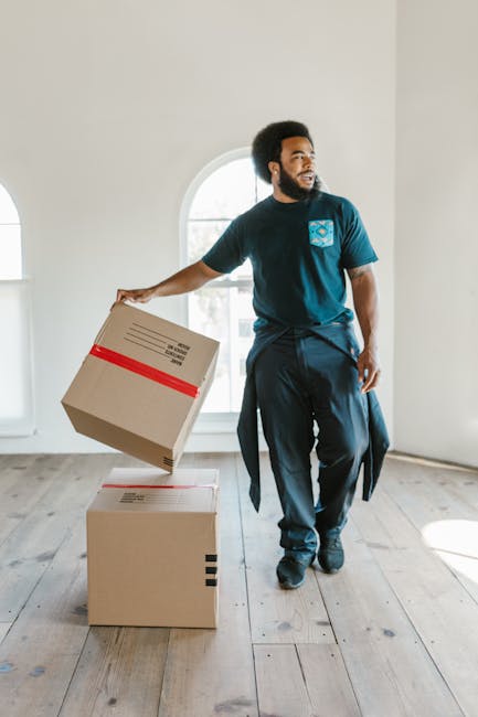 A man with dark curly hair and a beard, wearing a navy blue T-shirt and dark trousers, is shown inside a bright, spacious room with white walls and a large arched window that allows natural light to fill the space. He is holding a cardboard box with red tape around it in his right hand, while another similar cardboard box is positioned on the wooden floor nearby. The man appears to be in the process of loading or arranging moving boxes as part of a home relocation. The room is empty of furniture, and the floor is light-colored wood. This scene captures a moment of furniture transport or packing and moving preparations, typical of professional house removals carried out by companies like Movers Harringay in the context of a house or office move, reflecting the logistics involved in a typical furniture transport or packing process during a residential move.