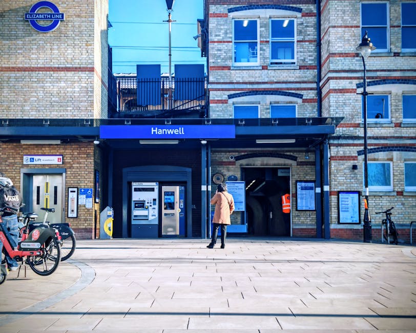 A view of Golders Green Underground Station with its elevated canopy featuring striped fabric awnings and white trim, under which several individuals are seen standing and sitting on wooden benches. Some figures are engaged in lifting or carrying cardboard boxes and duffle bags, indicating the process of home relocation or moving services. The scene captures a loading zone adjacent to the train tracks, with a yellow plastic container or trolley positioned nearby for transporting belongings. Beneath the canopy, an overhead sign directs towards the exit and indicates the location of lifts, while a CCTV camera is mounted on the station’s structure. Sunlight filters through, illuminating the area and highlighting the practical aspects of packing, loading, and transporting household or office items as part of a furniture transport or residential move, with the actions aligning with the services provided by Movers Harringay in the context of house removals.