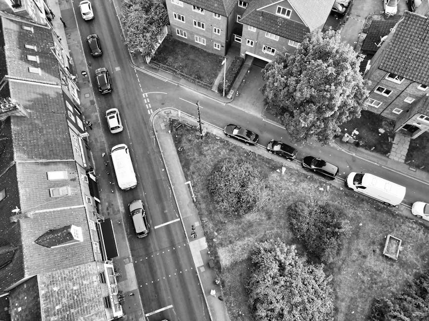 An aerial view captures a narrow residential street with a row of parked cars along the curb, including a white van positioned near the edge of the road, close to a small driveway leading to a housing complex. The street is partly obstructed by two large trees with dense foliage, and a patch of grass and bushes separates the road from the surrounding buildings. Some cars are parked parallel to the pavement, while others are parked at slight angles, indicating a busy or congested area. The scene shows the loading process of a home relocation or furniture transport, with the focus on the van, which appears to be ready for a moving service provided by Movers Harringay, suited for house removals and packing and moving logistics. The environment is well-lit, with light casting shadows from the vehicles and trees, highlighting the urban setting and the careful organization required during relocation activities, confirming compliance with parking rules for moving vans and permits as outlined in Haringey Council's regulations.