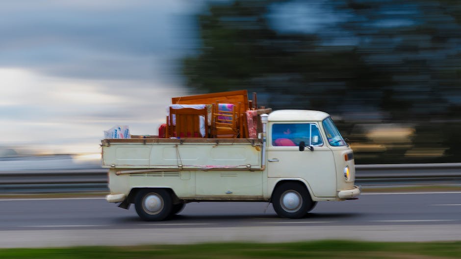 A vintage cream-colored small van is moving along a highway during daylight hours with a blurred background indicating motion. The van's flatbed area at the back is loaded with various household furniture and boxes, including a wooden chair, a small table, and several cardboard and plastic-wrapped items. The interior of the cab is partially visible through the windshield and side windows, showing a glimpse of the steering wheel and seating area. The van is part of a home relocation or furniture transport process, which may involve loading or unloading at a property. The environment suggests an outdoor setting with a guardrail and distant trees set against a cloudy sky. This image is associated with professional removals services, such as those offered by Movers Harringay, which assist with packing, moving, and transportation logistics, especially in compliance with local parking and permit regulations, as outlined in the Haringey Council parking rules for moving vans and permits, Harringay.