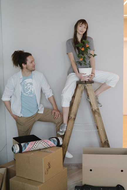 A young woman sits on a wooden step ladder, holding a potted plant with orange flowers and green foliage in a white container, while a young man stands nearby, looking at her. The man has curly dark h