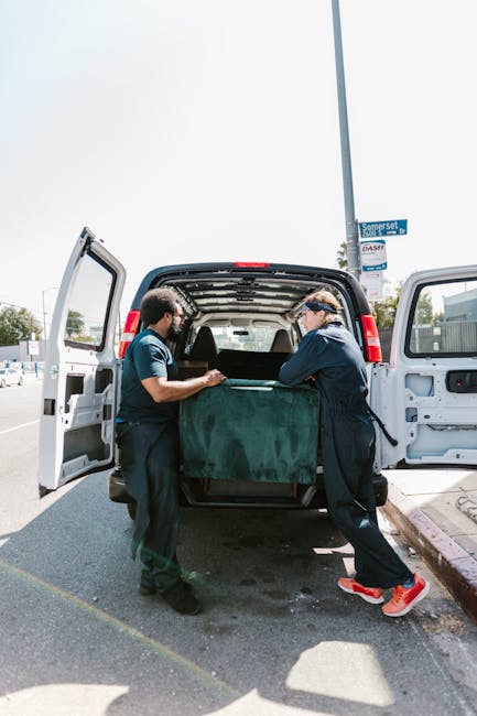 Two movers from Movers Harringay are seen loading a large, dark green piece of furniture into the rear cargo area of a black van during a home relocation. The van doors are wide open, revealing the interior space designed for furniture transport. The movers, dressed in dark work uniforms, are carefully handling the item, which is partially wrapped in protective material. The scene is set outdoors on a paved street, with a street sign visible in the background, indicating the location in Harringay. The surrounding environment includes parked cars, a lamppost, and some trees, under a clear sky with bright sunlight. This loading process involves the use of moving blankets and possibly straps or other equipment to secure items for transport, exemplifying professional packing and moving practices. Such activities are integral to efficient home removal services offered by Movers Harringay, ensuring careful handling during furniture transport and the packing and moving phases of a house relocation.