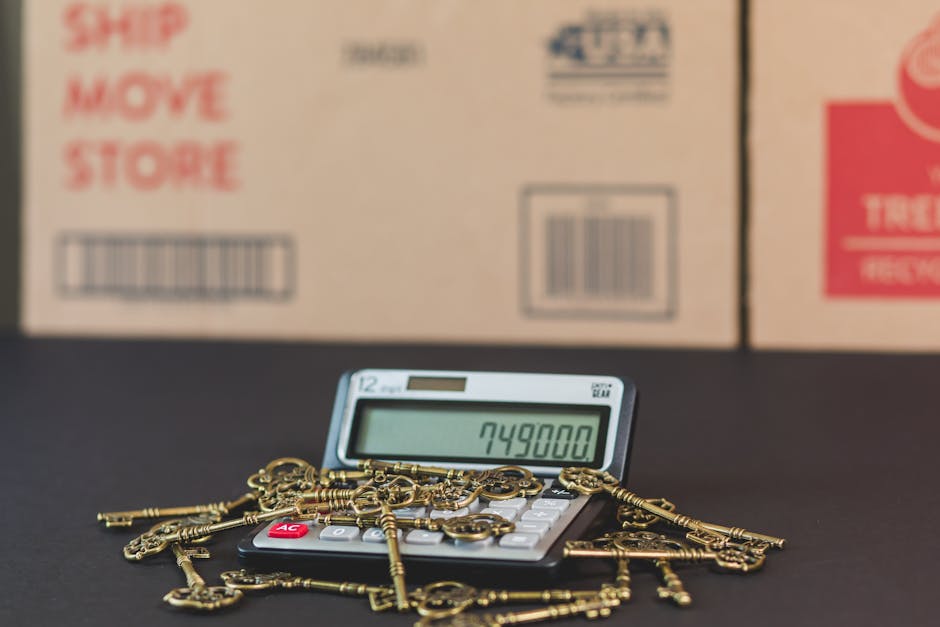 A small electronic calculator displaying the number 749800 is placed on a black surface, surrounded by a pile of vintage-style brass keys scattered across the electronic device. In the background, there are two cardboard boxes with red and black printed labels, indicating packaging materials for moving or storage. The setting appears to be indoors, with the boxes positioned close together, possibly inside a property prepared for a home relocation or packing process. The photograph captures the loading or packing stage of a house move, with the focus on the calculator and keys, subtly referencing the hidden costs to avoid in Harringay removal quotes, as offered by Movers Harringay's removals services.