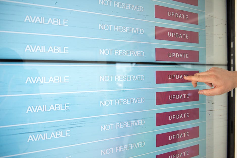 Inside a property, a person's hand is pointing towards a digital reservation screen displaying multiple rows of status information. The screen shows several labels, including 'AVAILABLE' in white text on a light blue background, and 'NOT RESERVED' in white text, alongside red buttons labeled 'UPDATE'. The environment appears to be well-lit, with reflections visible on the screen suggesting natural or bright interior lighting. This context is related to logistical planning for house removals or moving services, where real-time updates on availability or reservation status of moving slots or equipment are displayed. Occasionally, the digital display may be used by Movers Harringay for coordinating household furniture transport, packing, and loading processes during a home relocation, including managing the timing and scheduling of removals services.