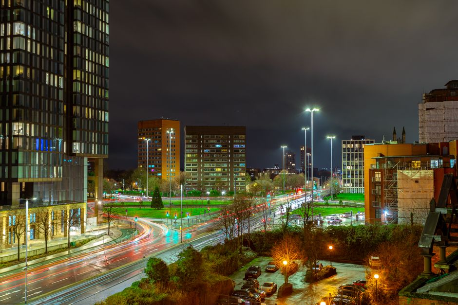 A nighttime cityscape showing a modern urban area during evening hours, with tall commercial and residential buildings illuminated by interior and exterior lights. In the foreground, there is a parking lot with several parked cars and surrounding trees, partially lit by streetlights. A main road runs through the scene, with visible light trails from moving vehicles indicating active traffic, likely captured with long exposure. To the right, a construction site with scaffolding and building materials is partially visible, suggesting ongoing development. Bright street lighting cascades down the road, highlighting the busy transportation and activity typical of a city preparing for home relocation or furniture transport. This scene was captured to illustrate the context of late evening removals and moving logistics in Harringay, with Movers Harringay providing services for packing, loading, and transport during extended hours.