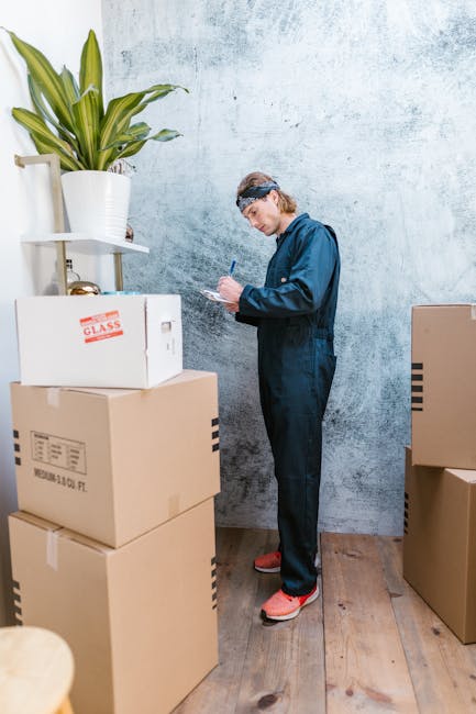 A person dressed in a navy blue jumpsuit and red shoes is standing indoors against a textured light blue wall, surrounded by multiple cardboard boxes used for home relocation. The individual is holdin