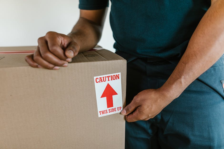 A person dressed in a navy blue jumpsuit and red shoes is standing indoors against a textured light blue wall, surrounded by multiple cardboard boxes used for home relocation. The individual is holdin